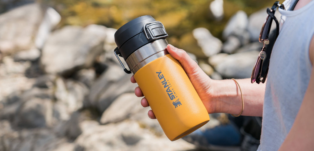 A person in a grey tank top drinking from a Stanley water bottle.